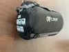 A black sleeping bag with gray straps and buckles lies on a wooden floor. Produktbild-Vorschau