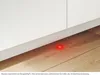 A close-up of the bottom of a white appliance on a wooden floor, with a red light illuminated on the floor. Produktbild-Vorschau