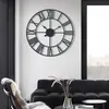 A large metal wall clock with Roman numerals hangs on a white wall above a black sofa and a chair with a fur throw. Produktbild-Vorschau