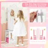 A young girl in a white dress is standing in front of a white jewelry cabinet with a mirror. The image is divided into sections, with one section showing a close-up of an anti-tip device being attached to a wall, and another section illustrating the stabil Náhľad obrázku produktu