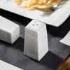 A close-up shot of two white ceramic salt and pepper shakers on a dark gray placemat, with a plate of french fries in the background. Produktbild-Vorschau