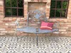 A grey metal garden bench with a decorative backrest sits on a patterned tiled patio in front of a brick wall with two small windows. Podgląd zdjęcia produktu