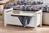 A white storage bench with a blanket and books inside, placed next to a bed with patterned bedding. Produktbild-Vorschau