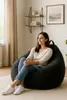 A woman is sitting comfortably in a large, dark gray beanbag chair in a well-lit room. The room has a minimalist decor with shelves on the wall and a framed picture. Voir les images du produit