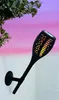 A black solar-powered garden torch with a flame effect is mounted on a white brick wall, with green leaves and a pink and purple sky in the background. Produktbild-Vorschau