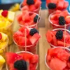 A close-up of several clear plastic cups filled with cut fruit, including watermelon, pineapple, and blackberries, arranged on a wooden surface. Náhľad obrázku produktu