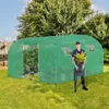 A man stands in front of a green greenhouse holding a potted plant. Náhled obrázku produktu
