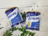 Two cans of Ensure vanilla flavored powder drink mix are displayed on a wooden surface with green leaves and white flowers. Produktbild-Vorschau