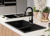 A modern kitchen sink with a black granite double basin and a black faucet, set on a white countertop with a built-in induction hob in the background. Decorative plants and a bowl of nuts are placed on the counter. Produktbild-Vorschau