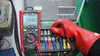 An electrician wearing red protective gloves uses a multimeter to test electrical connections on a panel. Produktbild-Vorschau