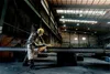 A worker in a factory is using an angle grinder on a metal beam, creating sparks. Podgląd zdjęcia produktu