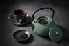 A cast iron teapot with a lid, a cup of tea, and loose tea leaves on a dark textured surface. Podgląd zdjęcia produktu