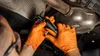 A person wearing orange gloves is working on the underside of a vehicle, holding a tool near the exhaust system. Produktbild-Vorschau