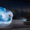 A living room with a large window overlooking a city skyline at night. A projector is set up on a round ottoman, projecting a dynamic image of a surfer riding a wave onto a wall. Anteprima dell’immagine prodotto