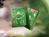 Two green playing card boxes with gold accents, one standing upright and the other tilted, against a soft, blurred background with hints of purple and green. Náhled obrázku produktu