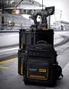 Tools and a backpack are displayed on a paved surface with a blurred background of a building and a racetrack. Podgląd zdjęcia produktu