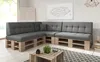 A sectional sofa made from wooden pallets with gray cushions, placed in a living room setting with a rug and a plant on a coffee table. Produktbild-Vorschau