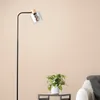 A modern floor lamp with a glass shade and wooden accents is placed next to a sofa with a blanket, with a small potted plant on a side table in the foreground. Produktbild-Vorschau