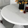 A close-up view of a white round table with a transparent protective film on top, showing a vase with yellow berries and a glass of dark liquid. Produktbild-Vorschau