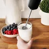 A hand is holding a glass of milk while a frother is being used to mix it. A bowl of berries is nearby. Produktbild-Vorschau