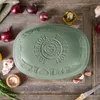 A green Römertopf casserole dish is placed on a rustic wooden table, surrounded by ingredients like red onions, garlic, rosemary, and a small bowl of salt. Produktbild-Vorschau