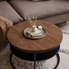A round coffee table with a wooden top and a black storage shelf underneath, placed on a patterned rug next to a sofa. Produktbild-Vorschau