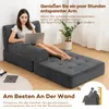 A woman is sitting on a dark gray sofa bed, using a laptop. The sofa is in a living room setting with a plant and a rug. Náhled obrázku produktu