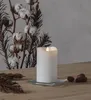 A white LED candle with a flickering flame sits on a glass dish on a wooden table, next to dried branches and cotton bolls. Podgląd zdjęcia produktu