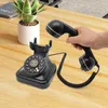 A hand holds the receiver of a vintage black rotary phone, which sits on a wooden desk next to a potted plant. Podgląd zdjęcia produktu
