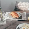 A croissant on a plate placed on a newspaper on a wooden table, with a vase of flowers and another plate with food in the foreground. Produktbild-Vorschau