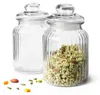 Two glass jars with lids, one filled with granola, on a white background with scattered seeds and nuts. Produktbild-Vorschau