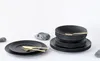 A set of black stoneware plates and bowls with gold cutlery, arranged on a white surface with decorative stones. Náhľad obrázku produktu