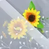 A close-up of a sheer white curtain with embroidered white leaf patterns and two artificial sunflowers. Produktbild-Vorschau