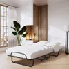 A foldable bed with a mattress in a room with a large window and plants. Voir les images du produit