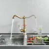 A vintage-style kitchen faucet is shown in use, with water running into a double sink. A rack of fresh produce is visible in the sink. Náhľad obrázku produktu