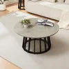 A round coffee table with a concrete-look top and a black metal base is placed on a light-colored rug in a living room setting. A small vase of flowers and a magazine with a cup of coffee are on the table. Podgląd zdjęcia produktu