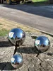 Three polished stainless steel garden balls are placed on a weathered tree stump, with a road and a yellow construction vehicle in the background. Produktbild-Vorschau