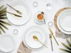 A flat lay of white dinnerware and gold cutlery on a white surface, with some greenery. Produktbild-Vorschau
