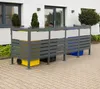A metal bin enclosure with three compartments, each containing a different colored trash bin, is situated on a paved patio in front of a modern building. Náhled obrázku produktu