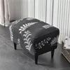 A dark gray ottoman with a white fern leaf pattern sits on a dark gray floor in front of a striped wall. A beige curtain is visible to the left. Produktbild-Vorschau