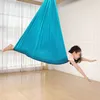 A young girl is using a blue aerial yoga hammock indoors, with her body extended and arms reaching forward. Náhled obrázku produktu