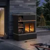 An outdoor fireplace on a wooden deck at dusk, with a basket of firewood and trees in the background. Náhled obrázku produktu