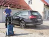 A man is washing a dark-colored station wagon with a pressure washer in front of a house. Produktbild-Vorschau