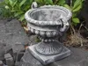 A stone amphora planter sits on rocks in a garden setting with greenery in the background. Produktbild-Vorschau