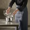 A person in an apron is lifting the lid off a steaming pot on a counter. Voir les images du produit