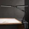 A black LED architect lamp with a dual head is clamped to a wooden desk, illuminating a large sheet of paper with drawings and two pencils. Náhľad obrázku produktu