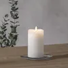 A white LED candle with a flickering flame sits on a square glass coaster on a wooden table. In the background, a grey wall is visible with a branch of eucalyptus leaves. Podgląd zdjęcia produktu