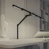 A black adjustable desk lamp with multiple light bars is positioned on a desk next to a white sculpture and an open book with a tablet. Náhled obrázku produktu