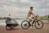 A man rides a bicycle with a child trailer attached, near a body of water. Produktbild-Vorschau
