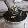 A round coffee table with a black glass top, a bouquet of flowers, a small bowl of candies, and a candle holder with candles. A grey sofa and a magazine are visible in the background. Produktbild-Vorschau